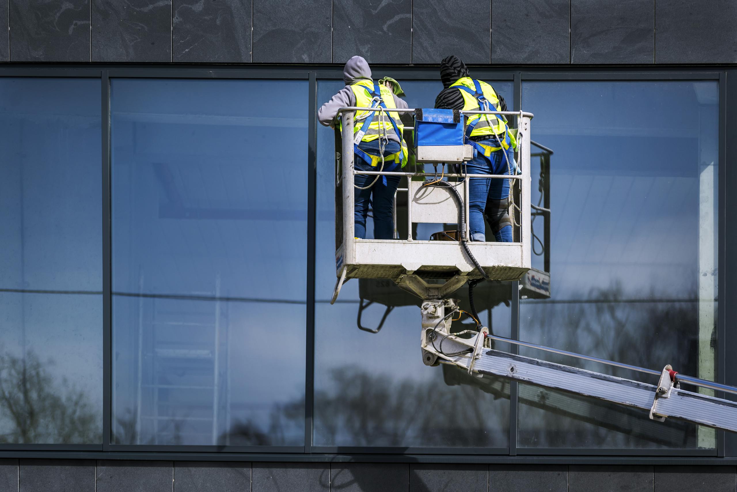 Two men on a hydraulic lift cleaning windows of a modern skyscraper. Urban reflection visible in the glass.